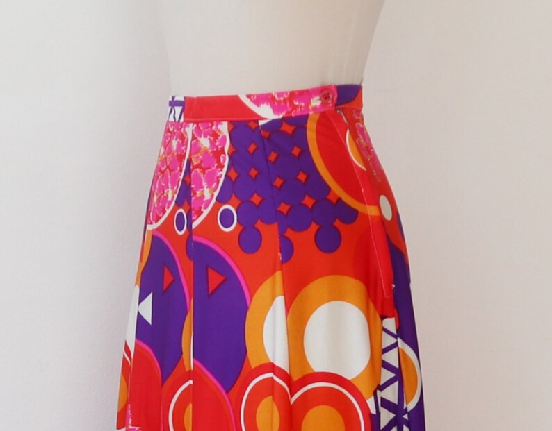 May include: A vibrant, high-waisted skirt featuring a bold, geometric pattern. The design incorporates large circles, triangles, and other shapes in shades of red, orange, purple, and white. The skirt has a button closure and a pleated design.