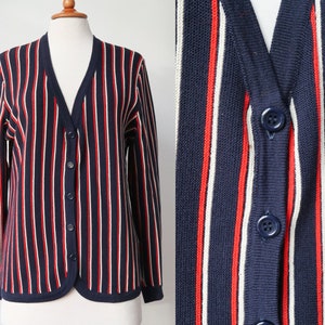 Op de afbeelding: Marineblau vest met rode en witte verticale strepen. De trui met knopen heeft een V-hals en lange mouwen. Het vest wordt op een mannequin gepresenteerd.
