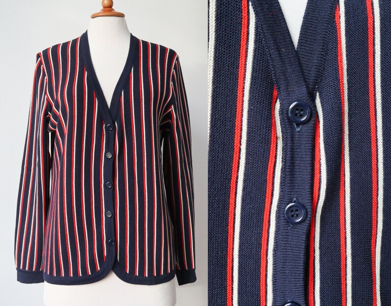Op de afbeelding: Marineblau vest met rode en witte verticale strepen. De trui met knopen heeft een V-hals en lange mouwen. Het vest wordt op een mannequin gepresenteerd.