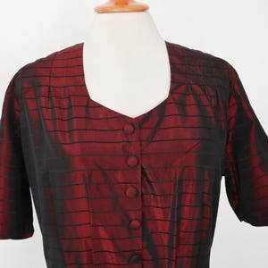 May include: A short-sleeved, button-down blouse in a deep red color with black horizontal stripes. The blouse has a square neckline and is displayed on a mannequin. The fabric has a subtle sheen, suggesting a silk or satin material.
