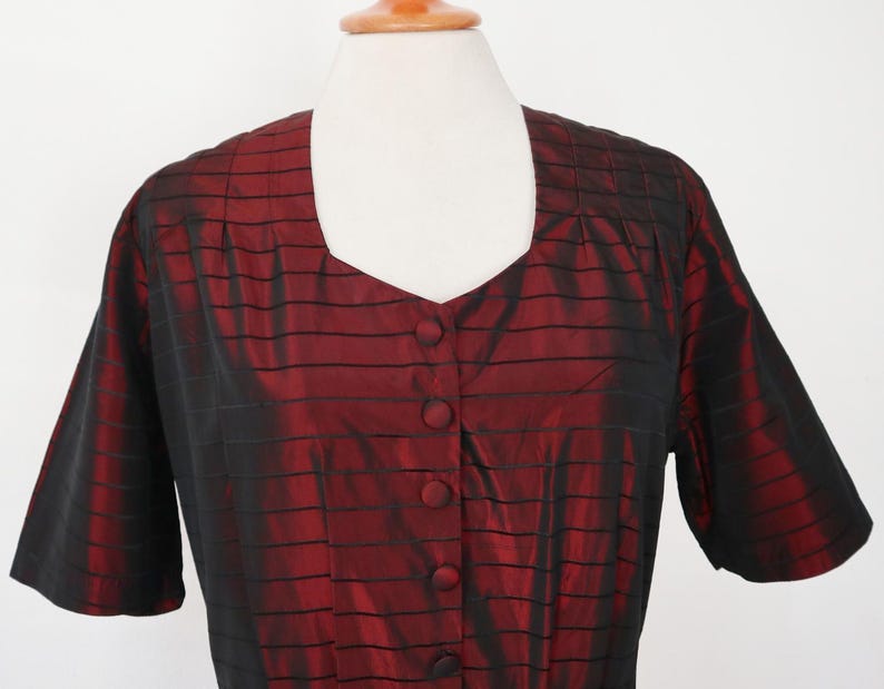 May include: A short-sleeved, button-down blouse in a deep red color with black horizontal stripes. The blouse has a square neckline and is displayed on a mannequin. The fabric has a subtle sheen, suggesting a silk or satin material.