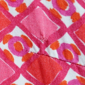 May include: Close-up of a fabric with a repeating pattern of pink, orange, and white geometric shapes. The design includes diamonds, circles, and other abstract forms, creating a vibrant and colorful textile.