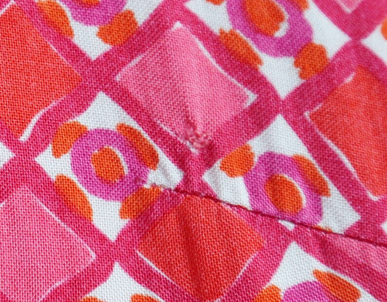 May include: Close-up of a fabric with a repeating pattern of pink, orange, and white geometric shapes. The design includes diamonds, circles, and other abstract forms, creating a vibrant and colorful textile.