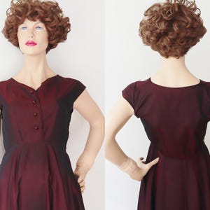 May include: A burgundy vintage dress with a V-neckline and cap sleeves. The dress features a button-down front and a flared skirt. The fabric has a subtle sheen, and the dress is displayed on a mannequin.
