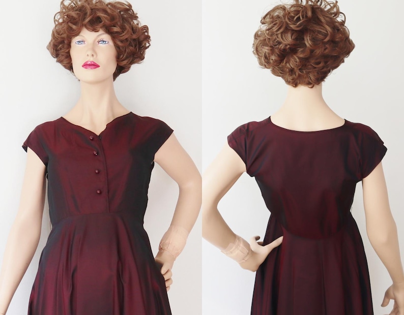 May include: A burgundy vintage dress with a V-neckline and cap sleeves. The dress features a button-down front and a flared skirt. The fabric has a subtle sheen, and the dress is displayed on a mannequin.