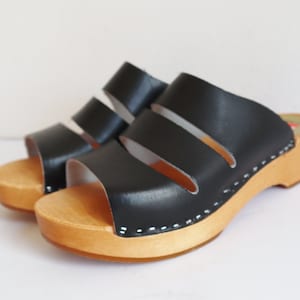 Puede incluir: Sandalias de cuero negro con suela de madera. Las sandalias tienen tres correas horizontales y una base de madera clara. La altura del tacón es de aproximadamente 7,6 cm.