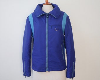 May include: A blue and light blue ski jacket with a full zipper closure. The jacket has a high collar, light blue accents on the shoulders, and a small pocket with a silver ring detail. The sleeves have zippers at the cuffs.