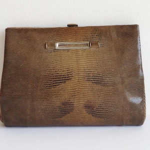 Pode incluir: Uma clutch marrom vintage com uma textura semelhante à de um réptil. A bolsa retangular apresenta um fecho prateado e uma barra decorativa. A clutch é provavelmente feita de couro ou material semelhante.