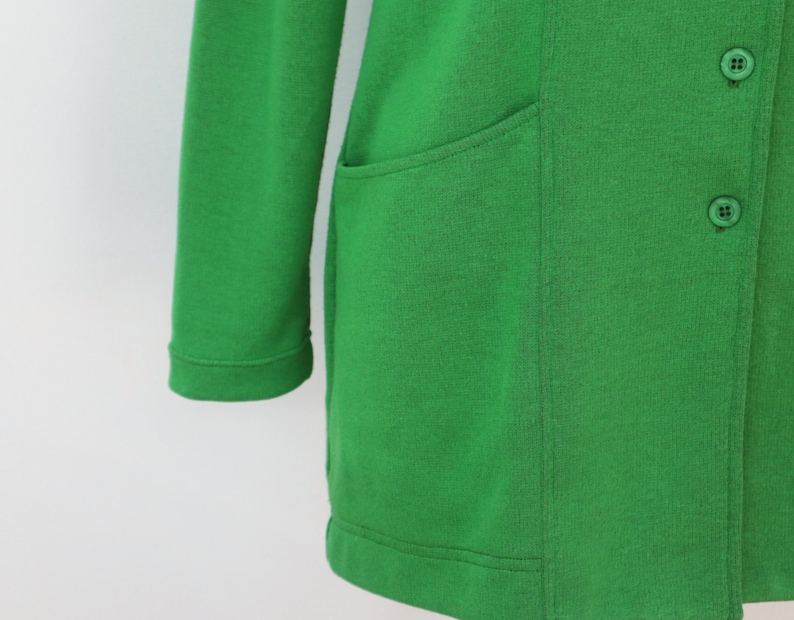 May include: A vibrant green long-sleeved cardigan featuring a smooth knit texture, a straight hemline, and two visible matching green buttons on the front placket. The garment includes a single patch pocket on the side with clean stitched seams.
