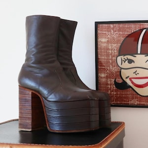 May include: A pair of brown leather platform boots with a tall shaft and wooden block heels. The boots have a stacked platform sole and are set on a black surface. A framed illustration of a woman wearing a helmet is in the background.
