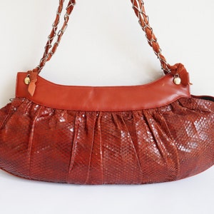 May include: A vintage, reddish-brown leather handbag with a snakeskin pattern. The bag features a wide, gathered body and a smooth, rectangular top in a darker shade. The straps are a combination of chain and suede.