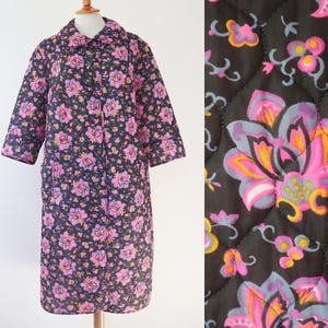 Könnte beinhalten: Ein Vintage-Steppmantel mit floralem Muster. Der Mantel hat einen schwarzen Hintergrund mit rosa, lila und gelben Blumen. Er hat einen Kragen, Dreiviertelärmel und Taschen. Der Mantel wird auf einer Holzpuppe präsentiert.