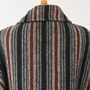 May include: A close-up of a wool coat with a striped pattern. The coat features vertical stripes in black, white, and rust-red. The collar is visible, and the coat is hanging on a wooden hanger.