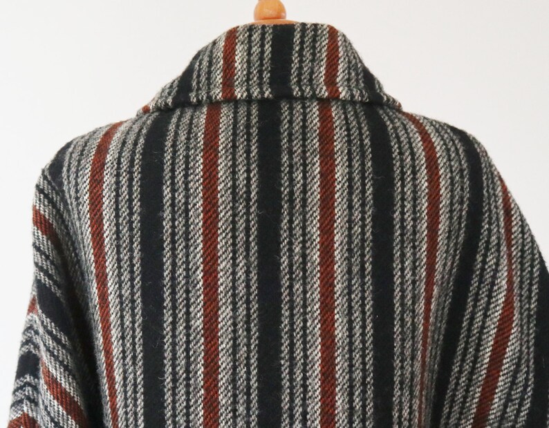 May include: A close-up of a wool coat with a striped pattern. The coat features vertical stripes in black, white, and rust-red. The collar is visible, and the coat is hanging on a wooden hanger.