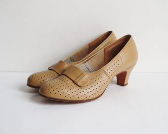 Op de afbeelding: Een paar bruinleren pumps met een geperforeerd ontwerp en een kleine hak. De schoenen hebben een ronde neus en een bandje over de bovenkant. De hakken zijn ongeveer 5 cm hoog.