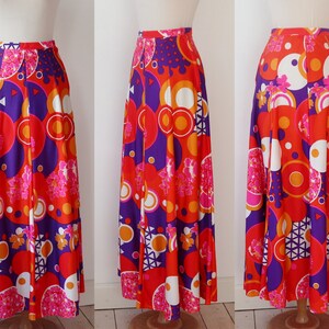 May include: A long, flowing skirt with a vibrant, retro-inspired print. The skirt features a bold pattern of red, orange, purple, white, and pink geometric shapes and floral accents. The skirt is displayed on a mannequin, showcasing its full length and design.