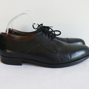 May include: A pair of black leather dress shoes with a cap toe design. The shoes have black laces and a low heel. The shoes are displayed on a white surface.