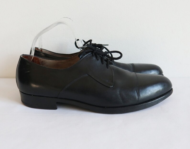 May include: A pair of black leather dress shoes with a cap toe design. The shoes have black laces and a low heel. The shoes are displayed on a white surface.