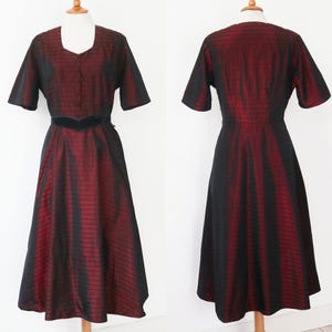 May include: A vintage midi dress in a striking red and black striped pattern. The dress features short sleeves, a square neckline, and a fitted bodice with a defined waist. The skirt flares out, creating an elegant silhouette. The dress is displayed on a mannequin.
