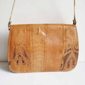 May include: A vintage, tan crossbody bag with a textured, reptile-like pattern. The bag has a flap closure and a long, adjustable strap. The design includes darker brown decorative accents on the front, adding visual interest to this classic accessory.