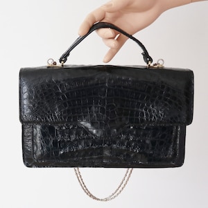 Può includere: Una borsa a mano nera con motivo testurizzato in pelle di coccodrillo, dotata di manico superiore e sottile tracolla a catena in metallo dorato. La borsa rettangolare presenta una chiusura a patta con anelli metallici dorati di fissaggio.
