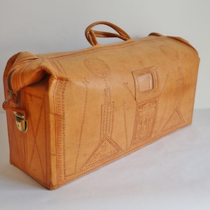 May include: A tan leather travel bag featuring intricate embossed geometric and architectural patterns across its surface. The bag includes a top handle, a rectangular identification window, and a gold-toned metal latch on the side zipper closure.
