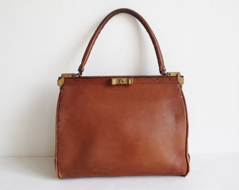 May include: A vintage brown leather handbag with a structured rectangular shape. The bag features a top handle, brass-colored metal accents, and a clasp closure. The leather shows signs of wear, adding to its antique charm.