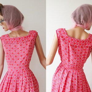 May include: A vintage sleeveless dress in shades of pink and white, featuring a geometric pattern. The dress has a square neckline and a pleated skirt. A pink scarf is draped over the head. The dress is displayed on a mannequin.
