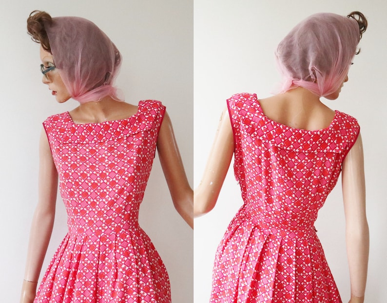 May include: A vintage sleeveless dress in shades of pink and white, featuring a geometric pattern. The dress has a square neckline and a pleated skirt. A pink scarf is draped over the head. The dress is displayed on a mannequin.