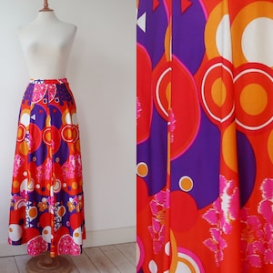 May include: A long, flowing skirt with a vibrant, retro design. The skirt features a bold pattern of large circles, geometric shapes, and floral accents in shades of red, orange, purple, and white. The skirt is displayed on a mannequin.