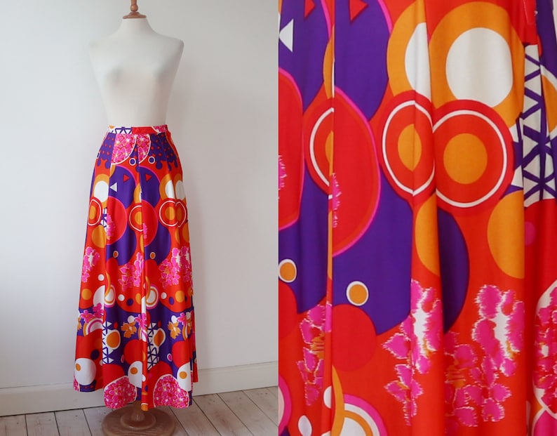 May include: A long, flowing skirt with a vibrant, retro design. The skirt features a bold pattern of large circles, geometric shapes, and floral accents in shades of red, orange, purple, and white. The skirt is displayed on a mannequin.