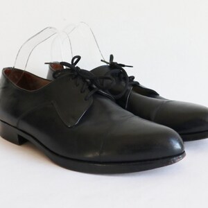 May include: A pair of black leather dress shoes with a classic cap-toe design. The shoes have a low heel and are laced up with black laces. The shoes are displayed on a white background.