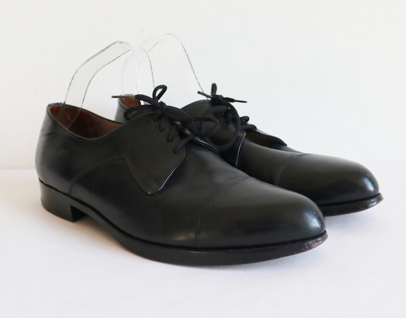 May include: A pair of black leather dress shoes with a classic cap-toe design. The shoes have a low heel and are laced up with black laces. The shoes are displayed on a white background.