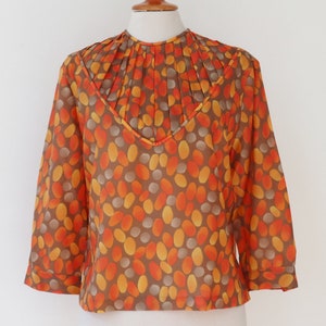 Op de afbeelding: Een vintage blouse met een bruine achtergrond en een patroon van oranje, gele en grijze ovale vormen. De blouse heeft een V-hals en driekwart mouwen. De halslijn heeft geplooid stof.