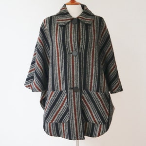 Op de afbeelding: Een vintage capejas met een gestreept patroon in zwart, grijs en bordeaux. De jas heeft een kraag, grote knopen en twee voorzakken. De cape heeft een relaxte pasvorm en driekwart mouwen.