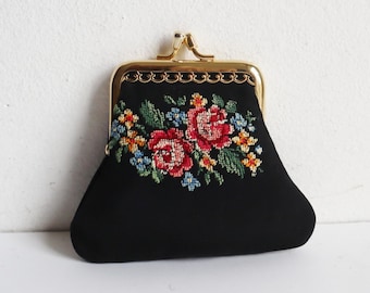 以下が含まれることがあります： 金色の留め金と花の刺繍が施された黒い小銭入れ。 刺繍には赤いバラ、青と黄色の花、緑の葉が描かれています。