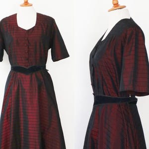 May include: A vintage-style dress in a two-piece design, featuring a striking combination of black and burgundy. The dress has short sleeves, a square neckline, and a fitted bodice with a button-down front. A black velvet belt cinches the waist.