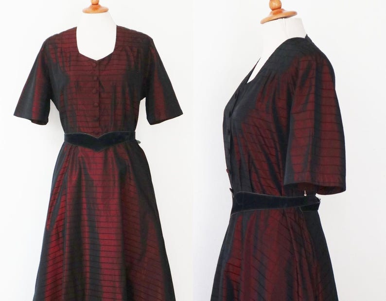 May include: A vintage-style dress in a two-piece design, featuring a striking combination of black and burgundy. The dress has short sleeves, a square neckline, and a fitted bodice with a button-down front. A black velvet belt cinches the waist.