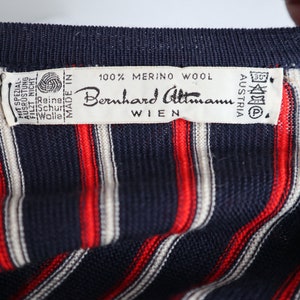 Op de afbeelding: Close-up van een marineblauw gebreid kledingstuk met verticale rode en witte strepen. Een label leest "100% Merino Wool" en "Bernhard Altmann Wien Austria". Het label bevat ook wasvoorschriften.