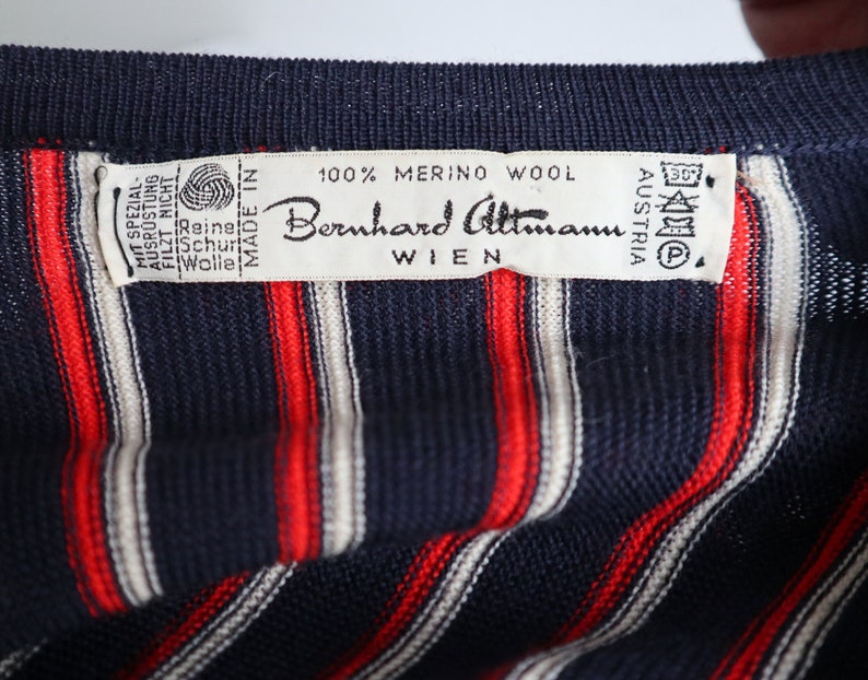 Op de afbeelding: Close-up van een marineblauw gebreid kledingstuk met verticale rode en witte strepen. Een label leest "100% Merino Wool" en "Bernhard Altmann Wien Austria". Het label bevat ook wasvoorschriften.