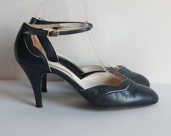 May include: A pair of navy blue high-heeled sandals with an ankle strap and a gold-colored buckle. The sandals have a pointed toe and a white trim along the edges. The heels are approximately 3 inches tall.