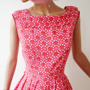 May include: A sleeveless dress with a square neckline. The dress features a repeating geometric pattern in shades of pink and red. The dress has a fitted bodice and a pleated skirt.
