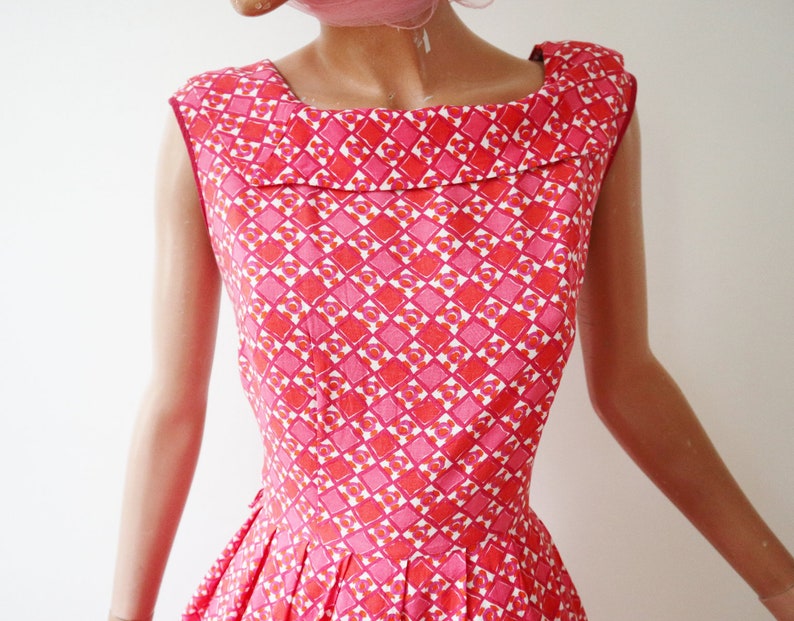 May include: A sleeveless dress with a square neckline. The dress features a repeating geometric pattern in shades of pink and red. The dress has a fitted bodice and a pleated skirt.