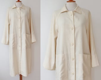 Puede incluir: Un abrigo color crema con botones, mangas largas y dos bolsillos delanteros. El abrigo se exhibe en un maniquí y tiene un cuello clásico. La tela parece ligera, adecuada para la primavera o el otoño.