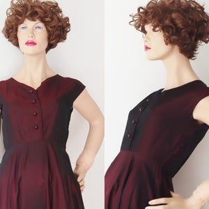 May include: A vintage burgundy dress with a black bodice and cap sleeves. The dress features a V-neckline and a row of buttons down the front. The skirt flares out from the waist, creating a classic silhouette. The dress is displayed on a mannequin.