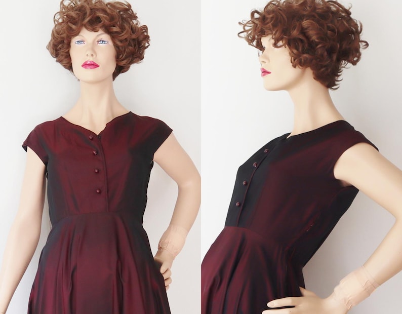 May include: A vintage burgundy dress with a black bodice and cap sleeves. The dress features a V-neckline and a row of buttons down the front. The skirt flares out from the waist, creating a classic silhouette. The dress is displayed on a mannequin.