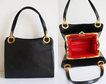 May include: Black leather handbag with gold-tone metal ring accents. The bag has a rectangular shape with a structured design. The interior is red, and the bag has a top handle and a clasp closure. The bag is a vintage style.