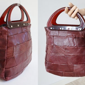 May include: A vintage burgundy leather tote bag with a patchwork design. The bag has a rectangular shape and a curved, dark brown handle. The handle is attached with silver rivets. The bag is suspended from a clear hook.