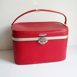 May include: A vintage red vanity case with a matching handle. The rectangular case has a silver-toned metal band and clasp. The word "antler" is visible on the case. The case is likely made of a textured material.