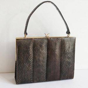 Peut inclure: Sac à main vintage marron avec un motif texturé rappelant la peau de reptile. Le sac rectangulaire est doté d'un fermoir doré et d'une poignée noire tressée. Le design comprend des panneaux verticaux.